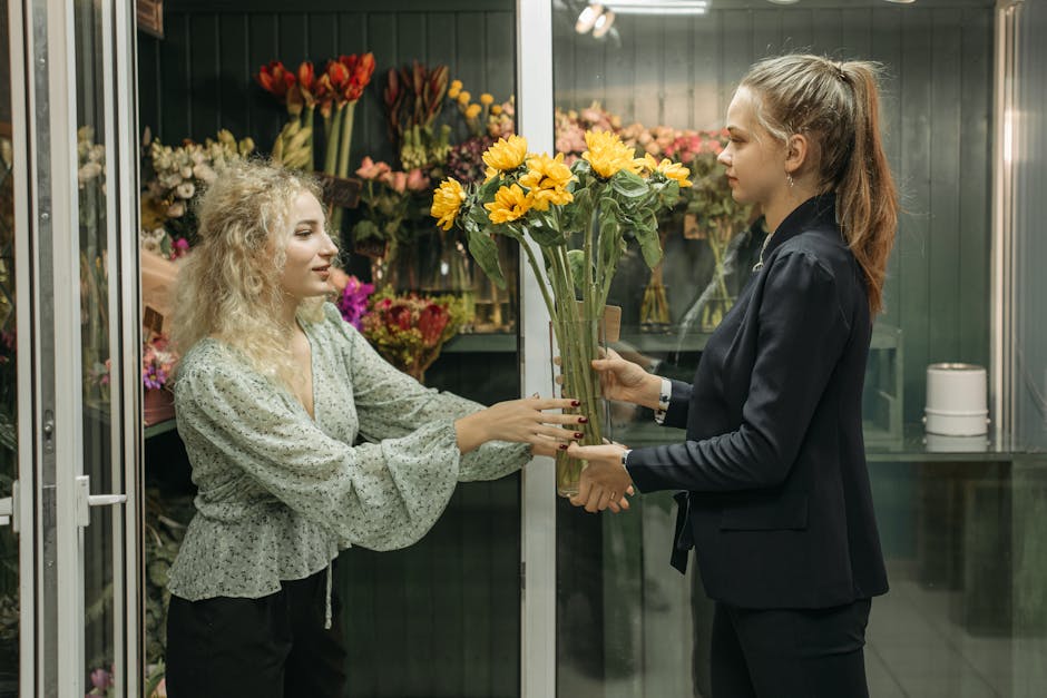 A florist in a dark blazer is handing a bouquet of bright yellow sunflowers to a customer with curly blonde hair wearing a light patterned blouse inside a floral shop. The bouquet is arranged with lon