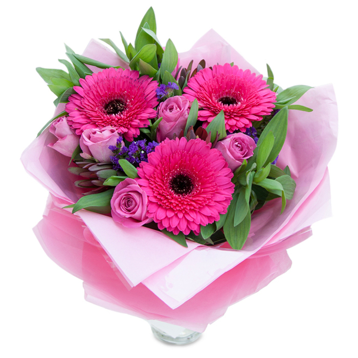 A charming, graceful bouquet that captures the playful yet refined spirit of spring in Bankside, presented on a crisp white backdrop that highlights its colour harmony. At its centre sit three exuberant fuchsia Gerbera daisies, their layered petals arcing out from deep, espresso-coloured centres that anchor the arrangement with bold contrast. Around them, lavender-pink roses unfurl slowly, their tight, velvety folds suggesting freshness and careful selection from cooler storage. Interspersed are delicate sprays of purple statice or limonium, tiny clustered blooms with a papery texture that add dusty-blue to violet notes and a meadow-like lightness. The foliage-broad, glossy leaves akin to salal and hints of eucalyptus-creates a verdant stage that amplifies the pinks while offering a crisp, slightly citrus-fresh scent. The bouquet is wrapped for gifting with an inner layer of soft, matte pale-pink tissue and an outer wrap of translucent, crinkled fuchsia-pink cellophane that catches studio light and gives the package a subtle sheen; the folds are neat, reflecting the hand of an experienced florist. The top rim of a clear glass vase shows at the bottom, indicating the arrangement is ready to be placed on a kitchen island or a Southbank windowsill overlooking the river. Texturally there is contrast between silky rose petals, the firmer flat leaves, and the papery statice; aromatically it is gentle-rose perfume softened by green notes. Ideal for birthdays, Mother's Day, anniversaries, or to lift a flat or workplace with a touch of local floral cheer.